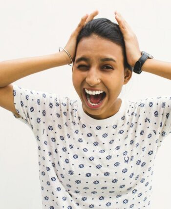 A young woman in a casual shirt screams with hands on her head against a white background.