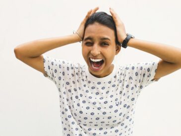 A young woman in a casual shirt screams with hands on her head against a white background.